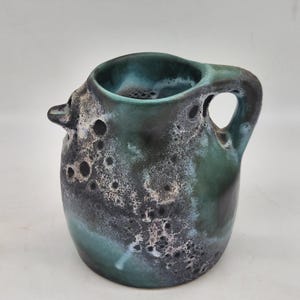 Peut inclure: Un pichet en céramique avec un émail tacheté dans des tons de bleu sarcelle, bleu foncé et noir. Le récipient présente un corps arrondi, un petit bec verseur et une anse incurvée. La texture de surface semble inégale avec des taches sombres.