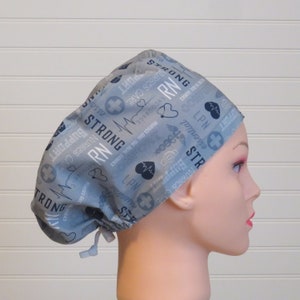 May include: A pale blue scrub cap featuring a repeating pattern of medical symbols, hearts, and text including "RN", "LPN", "Nurses Care", and "Strong". The cap has a drawstring closure and is designed for medical professionals.