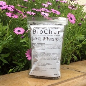 Może przedstawiać: Zbliżenie brązowego worka z nadrukiem "American Premium BioChar 100% Organic Fertilizer That Lasts A Lifetime" na przodzie. Worek leży na drewnianej powierzchni z r&oacute;żowymi kwiatami w tle.