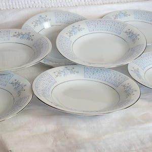 May include: A set of seven white ceramic soup bowls with a light blue floral pattern around the rim and a silver trim. The bowls are arranged on a white cloth.