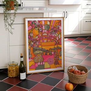 May include: A framed illustration of a colorful apple market with a pink and orange color scheme. The illustration is hanging on a white wall in a kitchen with black and red checkered tile flooring.