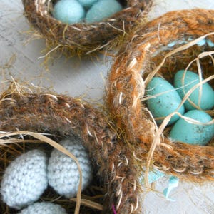 May include: Close-up of handmade decorative nests with blue eggs. The nests are crafted from brown and orange yarn, with straw accents. The eggs are a robin's egg blue color, creating a rustic and charming aesthetic. Ideal for Easter decor.