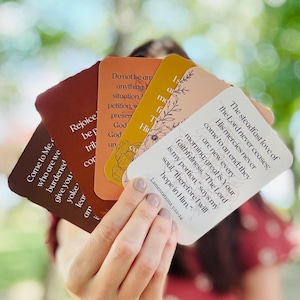 May include: A hand holds a fanned array of inspirational cards. Each card features a different color and design, with text from the Bible. The cards have rounded corners and are in shades of brown, orange, and white. Floral illustrations accent some cards.