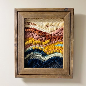 May include: A rectangular wall hanging featuring a hand-woven fiber art piece encased in a dark brown wooden frame. The textile displays horizontal layers of varying textures and colors, including cream, rust, pink, yellow, light blue, and navy blue.