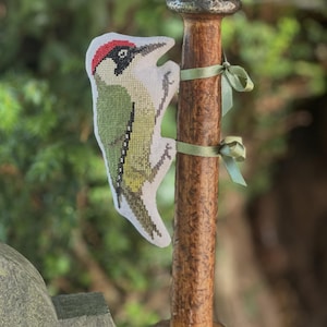 May include: A cross-stitched green woodpecker with a red crown and black eye patch, attached to a tall, cylindrical wooden spool with two light green ribbons. The spool has a dark, weathered texture and a flat, circular base.