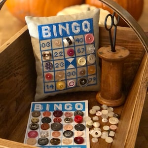 Puede incluir: Una bandeja de madera con una almohada beige que presenta un bordado de un cartón de bingo azul y blanco con botones. También hay un carrete de hilo, unas tijeras y un cartón de bingo con botones en la bandeja.
