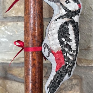 May include: A cross-stitched woodpecker ornament in black, white, and red thread, attached to a vertical wooden tool handle with two thin red satin ribbons. The woodpecker features a grey background fabric and a distinct red patch near its tail.