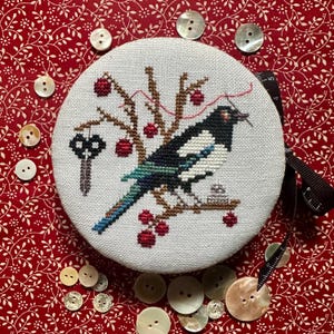 May include: A round, embroidered piece featuring a black and white magpie perched on a branch with red berries. A key hangs from the branch. The piece is surrounded by mother-of-pearl buttons and a black ribbon on a red floral background.