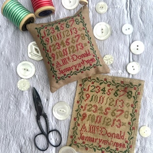 Op de afbeelding: Een stilleven met een kruissteek sampler en een klein kussen met geborduurde cijfers en tekst. Witte knopen, klosjes garen en een zwarte schaar omringen de sampler. De tekst luidt "A.M. McDonald. January 18th 1890."
