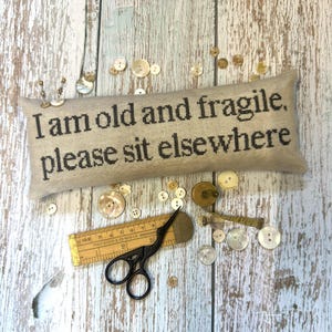 以下が含まれることがあります： 「I am old and fragile, please sit elsewhere」と黒字で書かれたベージュ色の長方形のクッション。様々なボタン、小さな定規、黒いハサミが、木製の表面にクッションの周りに散らばっています。