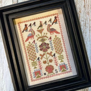 May include: A framed cross-stitch embroidery featuring a stylized floral design with birds and foliage in shades of red, green, blue, and brown. The embroidery is framed in a black frame with a double mat.
