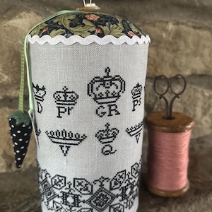 Könnte beinhalten: Ein zylindrisches Nadelkissen mit floral gemustertem Oberteil und einem gewellten weißen Besatz. Der graue Stoff ist mit schwarzen Kronen und geometrischen Mustern bestickt. Ein kleiner Erdbeer-Nadelhalter und eine Spule rosa Garn liegen daneben.