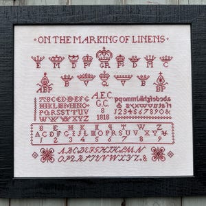 May include: Framed cross-stitch sampler with the text "ON THE MARKING OF LINENS" in red thread. The sampler features various crown designs, letters, numbers, and decorative elements, all stitched on a white background. The frame is black.