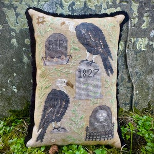 May include: A decorative pillow with a stitched design featuring two eagles perched on tombstones. The pillow has a beige background with black trim and a stitched design of tombstones and the letters "RIP".