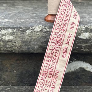 Peut inclure: Une bobine en bois entourée d'un ruban en lin beige orné de lettres de l'alphabet, de chiffres de 1 à 0 et du texte "Edith bone July 1884" brodés au point de croix rouge. Le ruban a une bordure rouge et un petit pompon rouge à l'extrémité.