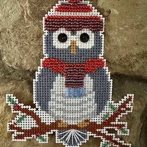 Pode incluir: Enfeite de coruja feito à mão em ponto cruz. A coruja usa um gorro e cachecol vermelhos, com olhos de botão pretos. Está pousada em um galho marrom com folhas verdes e bagas vermelhas. Uma decoração festiva com tema de inverno.