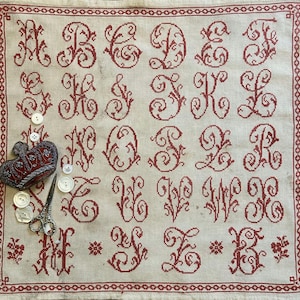 Puede incluir: Un muestrario de lino vintage con el alfabeto bordado en punto de cruz rojo. El muestrario tiene un borde rojo e incluye un alfiletero en forma de corazón, botones y tijeras. Las letras están en una fuente ornamental y decorativa.