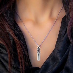 May include: A silver-colored pendant necklace featuring a cylindrical charm with a decorative filigree pattern and a small purple accent. The necklace is displayed against a dark background.