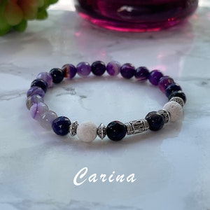 May include: A beaded bracelet featuring a mix of purple, black, and white stones. The bracelet includes a variety of bead shapes and textures, with a silver-toned metal accent. The word "Carina" is written in cursive on the bottom.