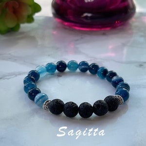 May include: A beaded bracelet featuring a gradient of blue and black beads. The bracelet includes light blue, dark blue, and black lava beads, with silver-colored spacers. The word "Sagitta" is printed in white.