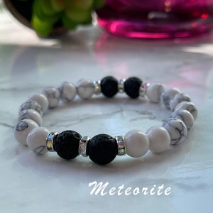 May include: A beaded bracelet featuring white and black lava stone beads. The white beads have a marbled pattern. The black beads are interspersed with clear, iridescent spacers. The word "Meteorite" is written in cursive at the bottom.