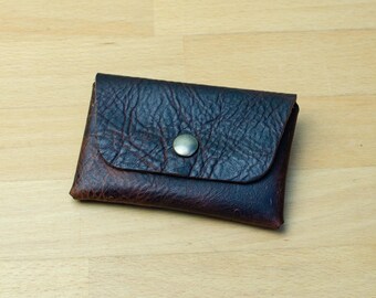 May include: A small, dark brown leather wallet with a textured surface and a flap secured by a silver-colored snap. The wallet is rectangular and appears to be empty. The background is a light-colored wooden surface.