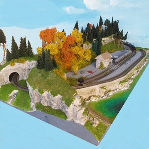 May include: A model train layout with a tunnel, a small building, and a winding track. The layout is covered in green grass and trees, including a large tree with orange and yellow leaves. The layout is set against a blue sky with white clouds.