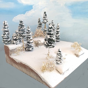 May include: A miniature winter landscape diorama featuring snow-covered evergreen trees and bushes. The scene is set on a snow-covered platform with a painted sky backdrop. The trees are dark green with white snow accents, creating a realistic winter scene.