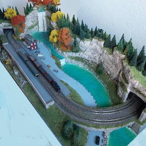 May include: A model train set with a train traveling through a tunnel and over a bridge. The train set is set up in a mountainous landscape with a waterfall and a river. The landscape is covered in green grass and trees.
