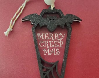 May include: Black glittery coffin-shaped ornament with a bat silhouette at the top and a spiderweb design at the bottom. The text "MERRY CREEP MAS" is in the center. The ornament hangs from a white and gold string.