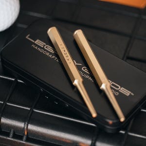 May include: Two gold-colored metal tools with pointed ends rest on a black box. The box has the text "LEGACY HANDCRAFTED EQUIPMENT" printed on it. A white golf ball is visible in the upper left corner.