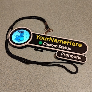 Puede incluir: Una placa de identificaci&oacute;n personalizada en blanco y negro con un icono circular que muestra un gr&aacute;fico de dinosaurio azul. La placa muestra el texto YourNameHere, Custom Status y Pronouns en letras blancas y amarillas, unida a un cord&oacute;n negro.