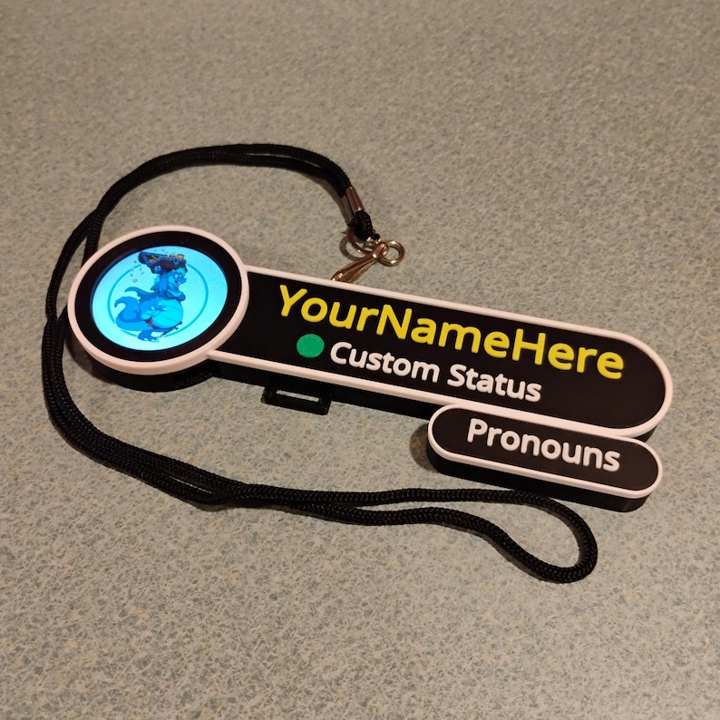 Puede incluir: Una placa de identificaci&oacute;n personalizada en blanco y negro con un icono circular que muestra un gr&aacute;fico de dinosaurio azul. La placa muestra el texto YourNameHere, Custom Status y Pronouns en letras blancas y amarillas, unida a un cord&oacute;n negro.