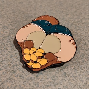 May include: A colourful enamel pin with a cartoon design. The pin shows a furry creature's backside in denim shorts, with a paw holding a cluster of yellow paw pads. The pin has a glossy finish and a black outline.