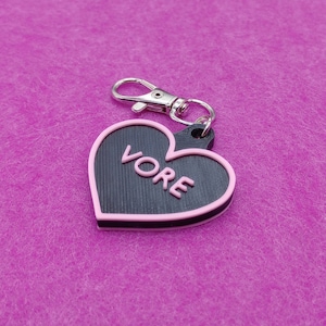 May include: A heart-shaped keychain with the word "VORE" in pink letters. The heart is black with a pink border and a silver-colored clasp. The keychain is likely made of plastic or a similar material.