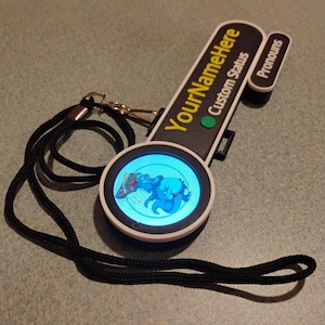 Puede incluir: Una insignia digital en blanco y negro con una pantalla circular que muestra un personaje de dibujos animados azul. La insignia presenta el texto YourNameHere, Custom Status y Pronouns, con un bot&oacute;n verde y un cord&oacute;n negro.