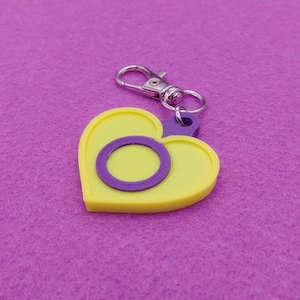 May include: A yellow heart-shaped keychain with a purple ring and a purple attachment. The keychain has a silver clasp for easy attachment to keys or bags. The design is simple and colorful.