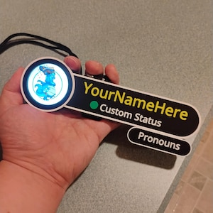 Puede incluir: Una insignia personalizada con luz LED circular y un gr&aacute;fico de drag&oacute;n azul. La extensi&oacute;n rectangular negra muestra el texto amarillo YourNameHere, un punto verde, el texto blanco Custom Status y una etiqueta inferior con la palabra Pronouns.