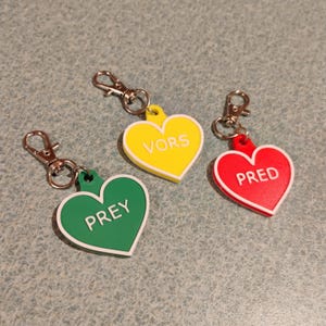 May include: Three heart-shaped keychains in yellow, green, and red. Each keychain has a white border and a silver clasp. The yellow keychain reads "VORS", the green keychain reads "PREY", and the red keychain reads "PRED".