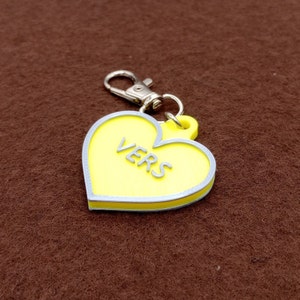 Puede incluir: Un llavero amarillo en forma de corazón con la palabra "VERS" en letras plateadas. El llavero tiene un cierre plateado y un borde plateado alrededor del corazón. El fondo es de color marrón liso.