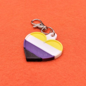 May include: A heart-shaped keyring displaying the non-binary pride flag colours: yellow, white, purple, and black. The keyring has a silver-coloured clasp and a white loop. The background is a solid orange colour.