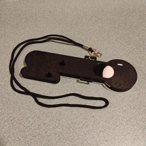 Puede incluir: Un soporte impreso en 3D en blanco y negro para un dispositivo de seguimiento circular, con un cuerpo rectangular largo y un extremo redondeado. El soporte incluye un cord&oacute;n negro sujeto por un cierre giratorio de metal para llevarlo.