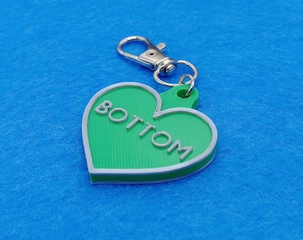May include: A green heart-shaped keychain with the word "BOTTOM" in white lettering. The keychain has a silver clasp and a white border. The background is a solid blue color.