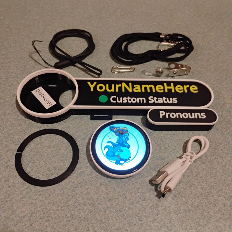 Puede incluir: Un conjunto de placa de identificaci&oacute;n personalizada con marco blanco y negro con el texto YourNameHere y Custom Status, una etiqueta Pronouns, una pantalla digital circular con un drag&oacute;n azul, un cord&oacute;n negro, cable y un anillo.