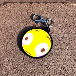 May include: A yellow, round keychain with a cartoon face design. The face has large white eyes with brown pupils, pink accents, and brown curved lines. The keychain has a black border and a silver clasp.