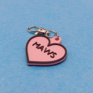 May include: A pink heart-shaped keychain with the word "MAWS" in black lettering. The keychain has a black border and a silver-colored clasp. The background is a light blue color.