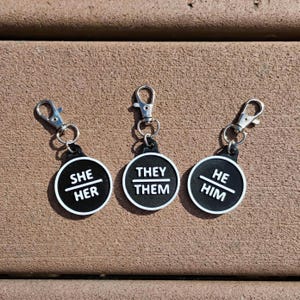 Może przedstawiać: Trzy breloczki do kluczy z czarno-białymi etykietami z zaimkami i srebrnymi zapięciami. Każda etykieta jest okrągła z białą obwódką. Etykiety wyświetlają zaimki "She/Her", "They/Them" i "He/Him" w białym tekście.