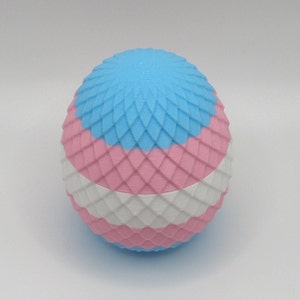 Op de afbeelding: Een 3D-geprint, bolvormig object met een getextureerd oppervlak. De bol is verdeeld in horizontale banden van blauw, roze en wit, die op een vlag lijken. Het ontwerp heeft een herhalend ruitpatroon over het oppervlak.