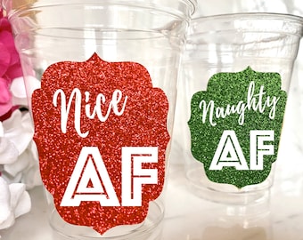 May include: Two clear plastic cups with glittery labels. One label is red and says "Nice AF", the other is green and says "Naughty AF". The cups are likely for a holiday or party.