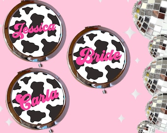 May include: Three round, silver-framed compact mirrors with black and white cow print designs. The mirrors have pink lettering that reads "Jessica", "Bride", and "Carla".